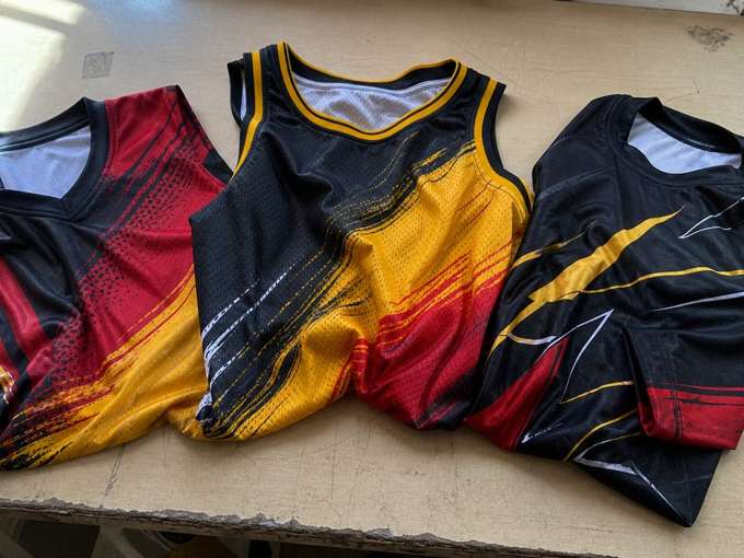 Tres prendas deportivas sublimadas sobre la mesa de corte del taller: jersey de cuello en V, camiseta de tirantes de básquet y camiseta de manga larga